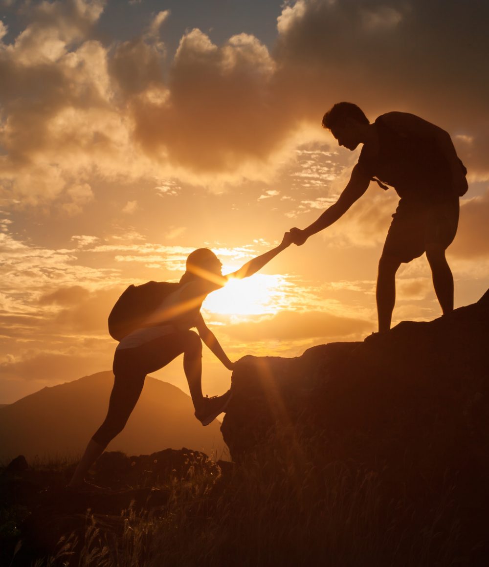 Male and female hikers climbing up mountain cliff and one of them giving helping hand. People helping and, team work concept.